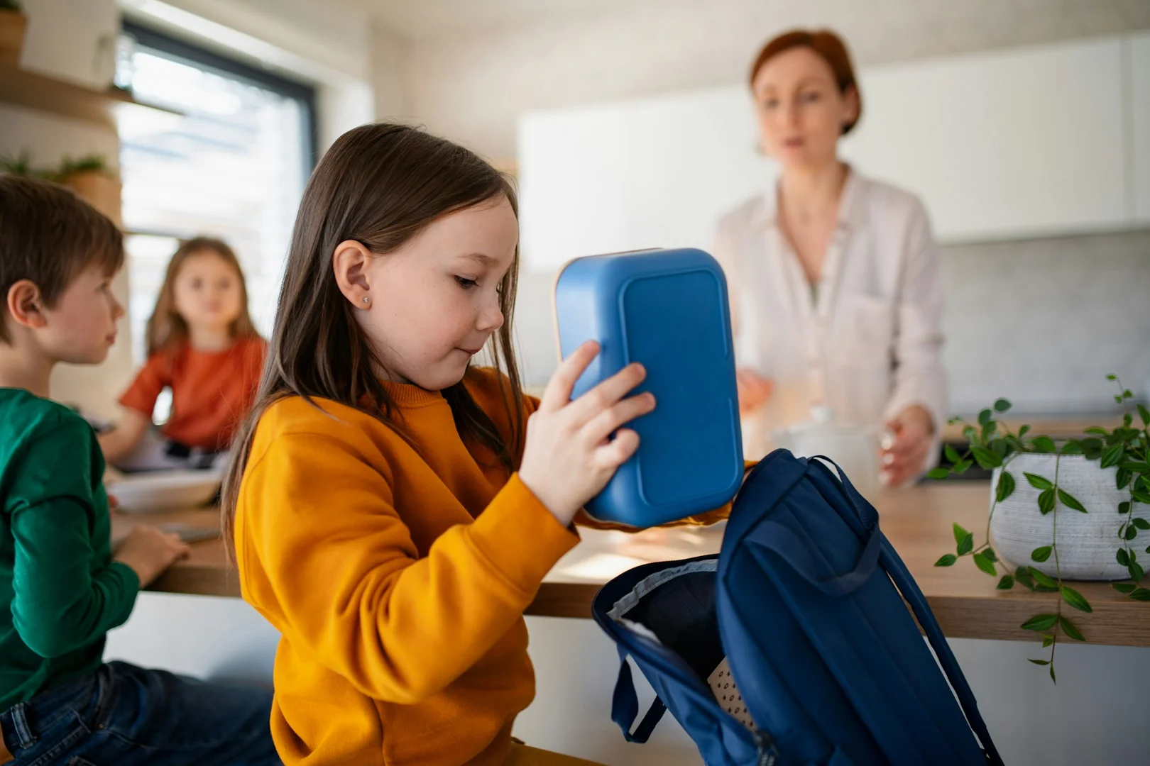 A young child packing a lunch box at home as a parent prepares breakfast in the background.